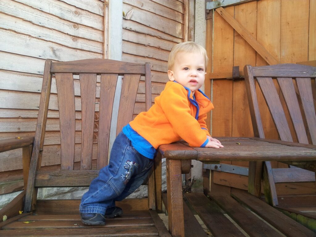 A toddler wearing a bright orange jacket and blue jeans stands on a wooden chair, leaning forward with hands on a table. The background shows a wooden fence and another empty chair. The child looks slightly to the right with a neutral expression.