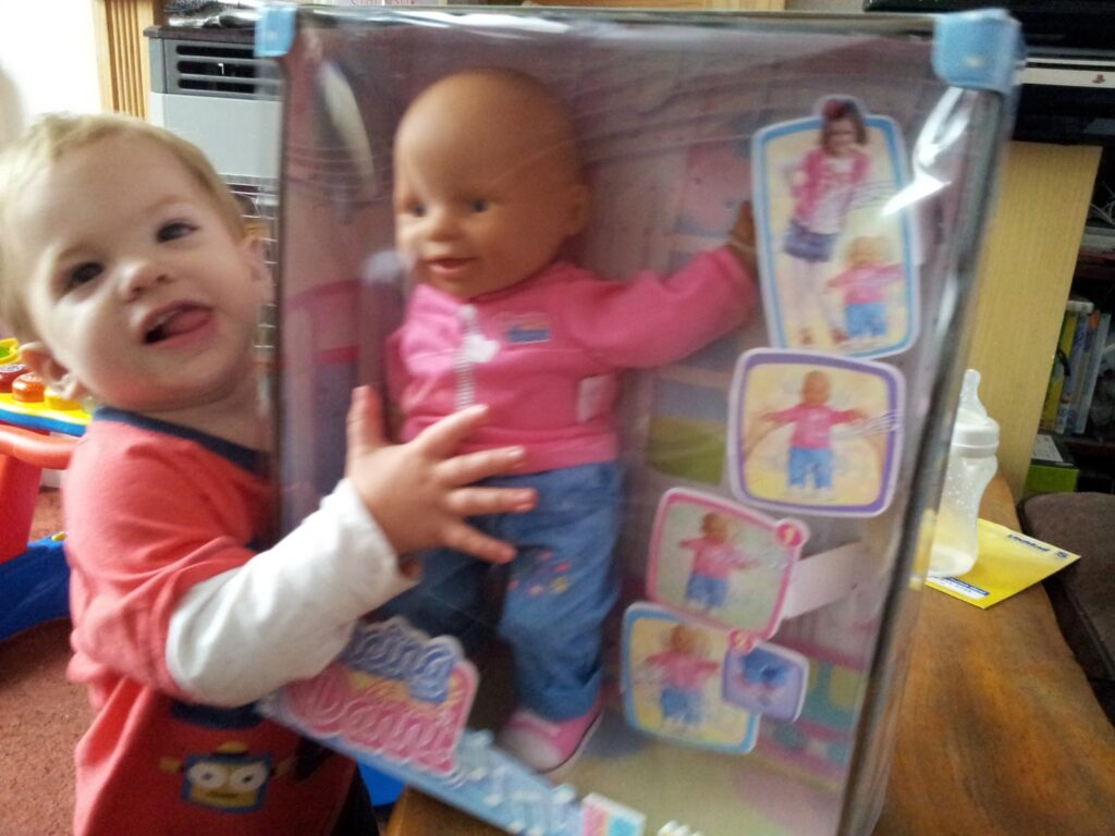 A smiling toddler with blonde hair wears a red shirt and hugs a boxed baby doll toy. The doll is dressed in a pink jacket and blue trousers. The background shows a bottle, papers, and toys on a table and shelves.