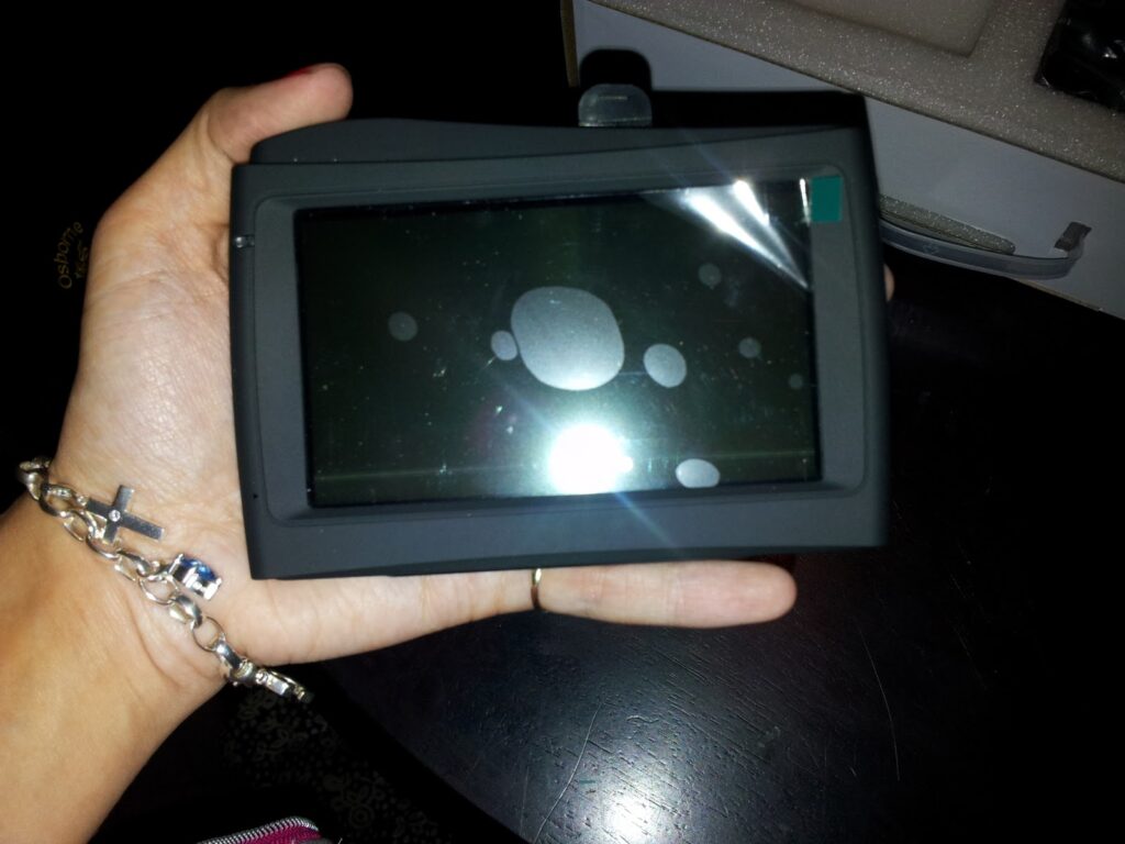 A hand holding a black touchscreen device with a reflective screen, showing several bubble marks and a glare, likely from a camera flash. The person wears a silver bracelet and a thin ring, and the background includes a dark table and part of a box.