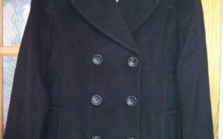 A black, double-breasted pea coat with wide lapels is hanging on a hanger. The coat features six visible buttons in two rows on the front and has slanted pockets. It hangs on a wooden door with frosted glass panes.