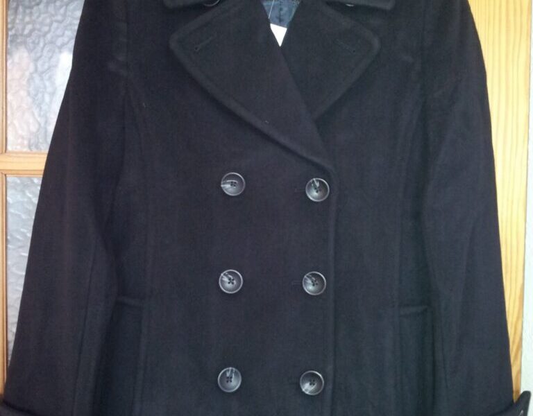 A black, double-breasted pea coat with wide lapels is hanging on a hanger. The coat features six visible buttons in two rows on the front and has slanted pockets. It hangs on a wooden door with frosted glass panes.