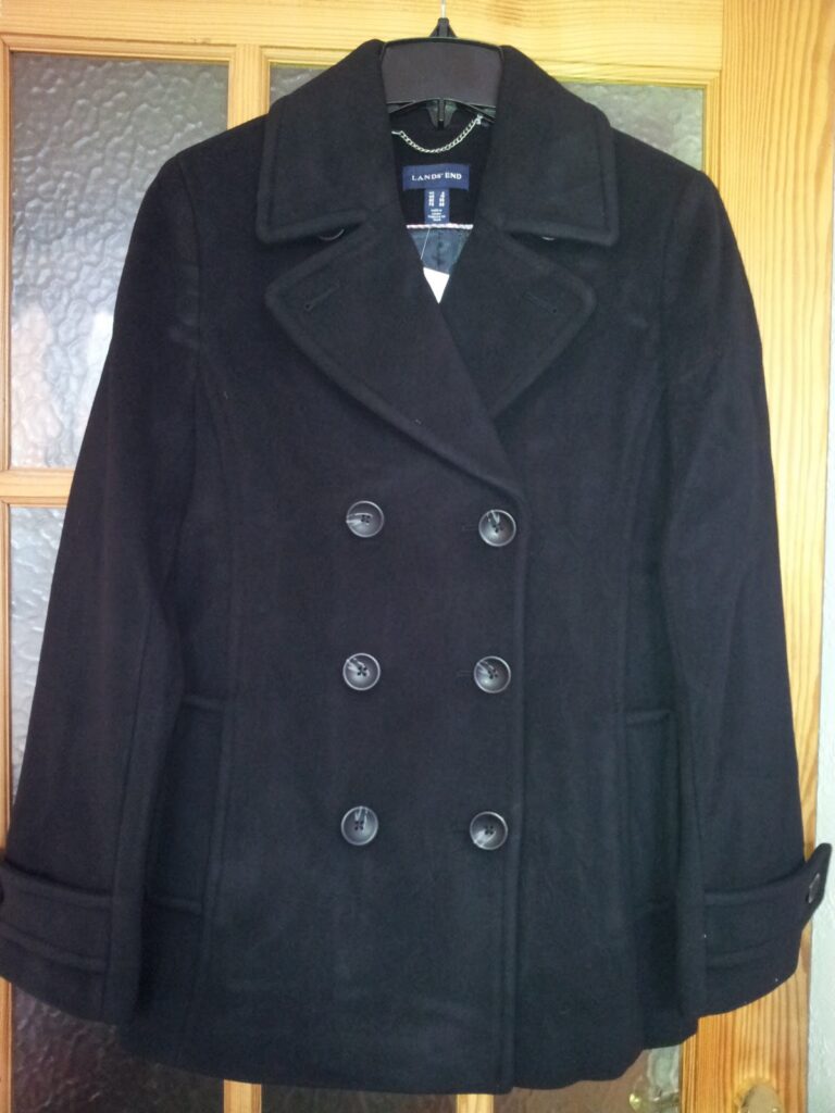 A classic black pea coat with a double-breasted front featuring six black buttons, wide notch lapels, and two front pockets, hanging on a wooden hanger in front of a wooden door with frosted glass panes.