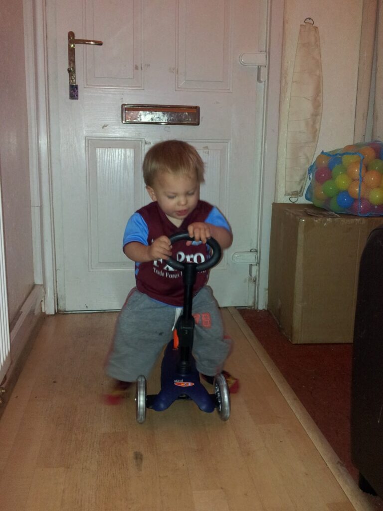 A young child wearing a sports jersey and grey trousers rides a blue and black three-wheeled scooter indoors on a wooden floor. Behind the child is a white door, a box filled with colourful plastic balls, and a beige folded item against the wall.