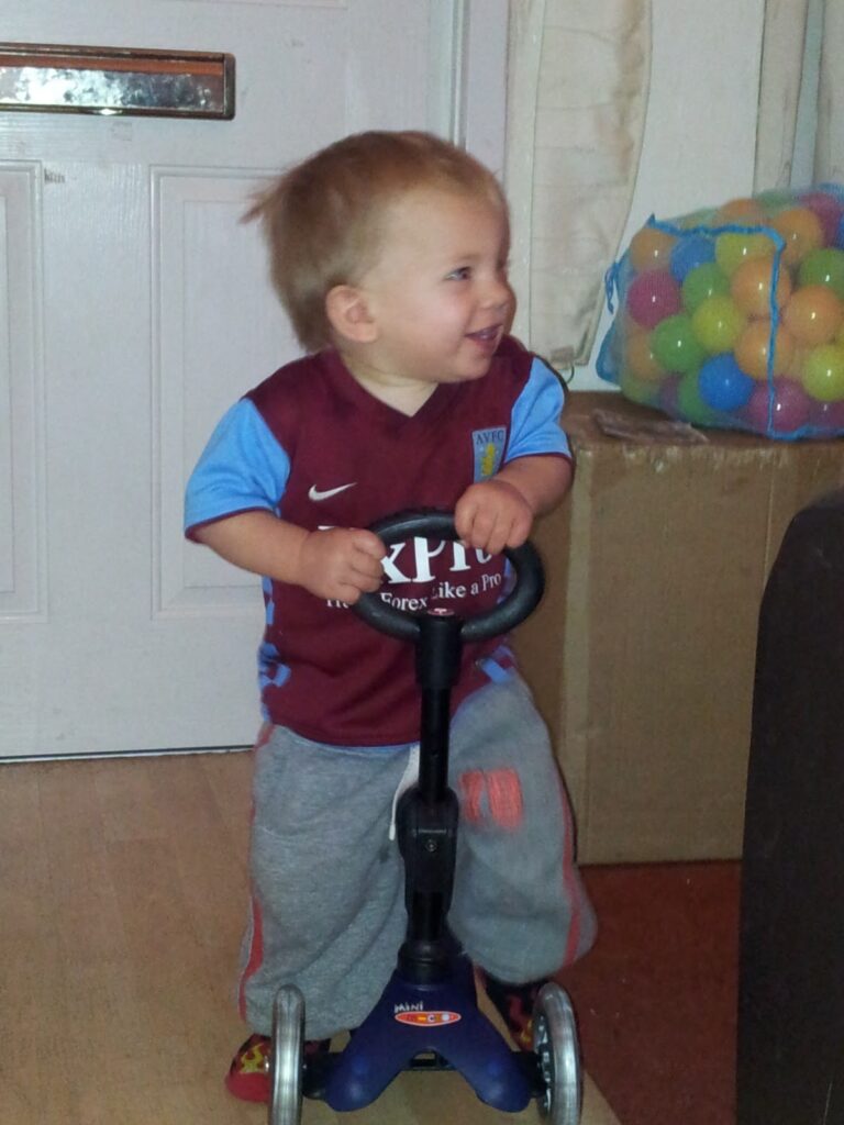 A toddler in a maroon and blue football shirt and grey trousers stands indoors, smiling while holding the handlebars of a small blue scooter. Behind the child is a box filled with colourful plastic balls and a white door with a letterbox.