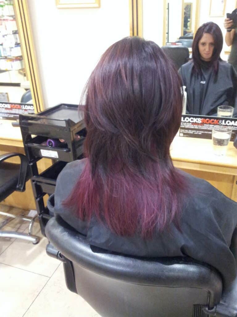 A person with layered, shoulder-length hair sits in a salon chair facing a mirror. Their dark hair transitions to a purple ombre at the ends. The salon has light-coloured walls, shelves with products, and a stylist is partially visible in the mirror.