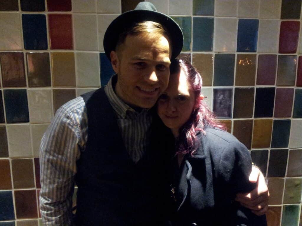 A man in a hat and striped shirt stands arm-in-arm with a woman in a dark coat. They are smiling and standing close together in front of a wall made of multicoloured square tiles. The lighting is dim, casting shadows on their faces.