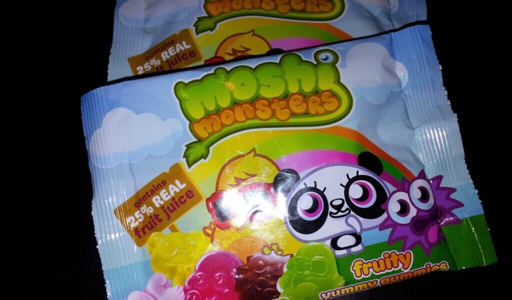 Two packets of Moshi Monsters fruity gummy sweets, featuring colourful cartoon characters and images of gummy bears on the front. The packaging highlights “25% real fruit juice,” “No artificial,” and has a blue sky with clouds and rainbows background.