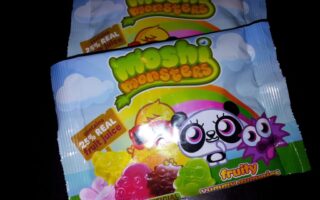 Two packets of Moshi Monsters fruity gummy sweets, featuring colourful cartoon characters and images of gummy bears on the front. The packaging highlights “25% real fruit juice,” “No artificial,” and has a blue sky with clouds and rainbows background.