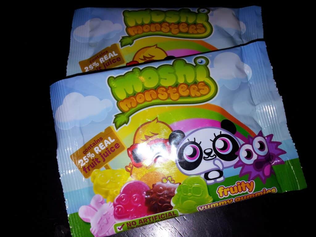 Two colourful packets of “Moshi Monsters” fruity gummy sweets are stacked on a dark surface. The packaging features cartoon monster characters, a rainbow, clouds, and mentions “25% real fruit juice” and “NO ARTIFICIAL” ingredients. Sweets are shown at the bottom.