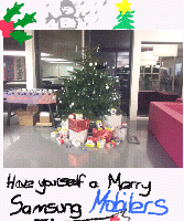 A decorated Christmas tree stands in an office, surrounded by wrapped presents. At the top, there are hand-drawn festive doodles: a snowman, holly, and a tree. Text below reads: "Have yourself a Merry Samsung Mobiles Christmas.