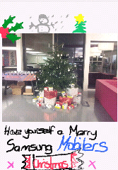 A decorated Christmas tree stands in an office, surrounded by wrapped presents. At the top, there are hand-drawn festive doodles: a snowman, holly, and a tree. Text below reads: "Have yourself a Merry Samsung Mobiles Christmas.