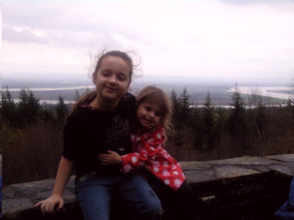 Two young girls sit on a stone wall overlooking a forested landscape with rivers and cloudy skies in the background. The older girl wears a dark jumper, while the younger, who is smiling, wears a red polka-dot coat. Both have windswept hair.