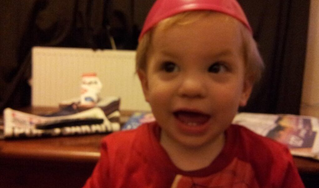 A young child with blond hair smiles widely, wearing a red shirt and a pink plastic bowl upside-down on their head. The background includes a table with newspapers and a small snowman decoration, with dark curtains and a white radiator behind.