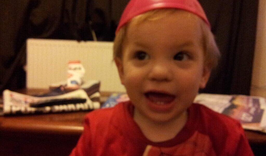 A young child with blond hair smiles widely, wearing a red shirt and a pink plastic bowl upside-down on their head. The background includes a table with newspapers and a small snowman decoration, with dark curtains and a white radiator behind.