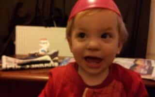 A young child with blond hair smiles widely, wearing a red shirt and a pink plastic bowl upside-down on their head. The background includes a table with newspapers and a small snowman decoration, with dark curtains and a white radiator behind.