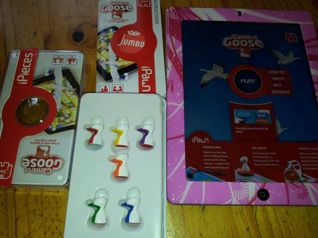 A wooden table holds a Game of Goose iPad board game set, including an open iPieces box, a Jumbo-branded token tray with six colourful goose pawns, and an iPad displaying the Game of Goose app’s menu screen.