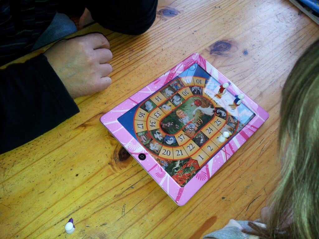 Two people are playing a board game with a pink, rectangular board featuring numbered squares in a spiral pattern and a central image. Small white pieces are on the board, which sits on a wooden table. Only the players' arms and hands are visible.