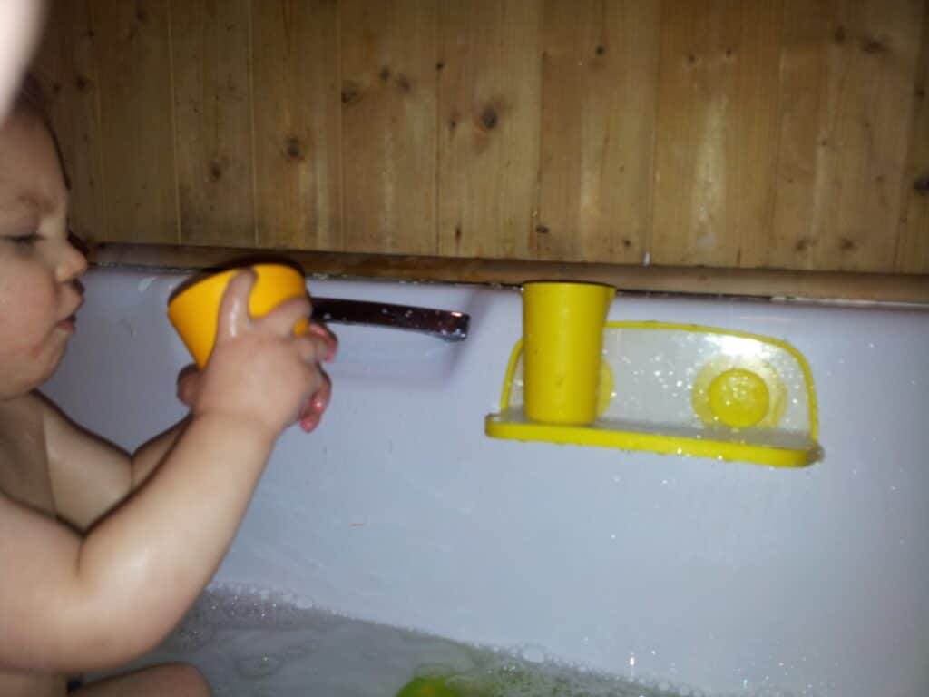 A young child sits in a bath, holding a yellow cup with both hands. Another yellow cup rests on a yellow holder attached to the bath wall. The bath has bubbles and the wall behind is wooden. Only the child’s upper body is visible.