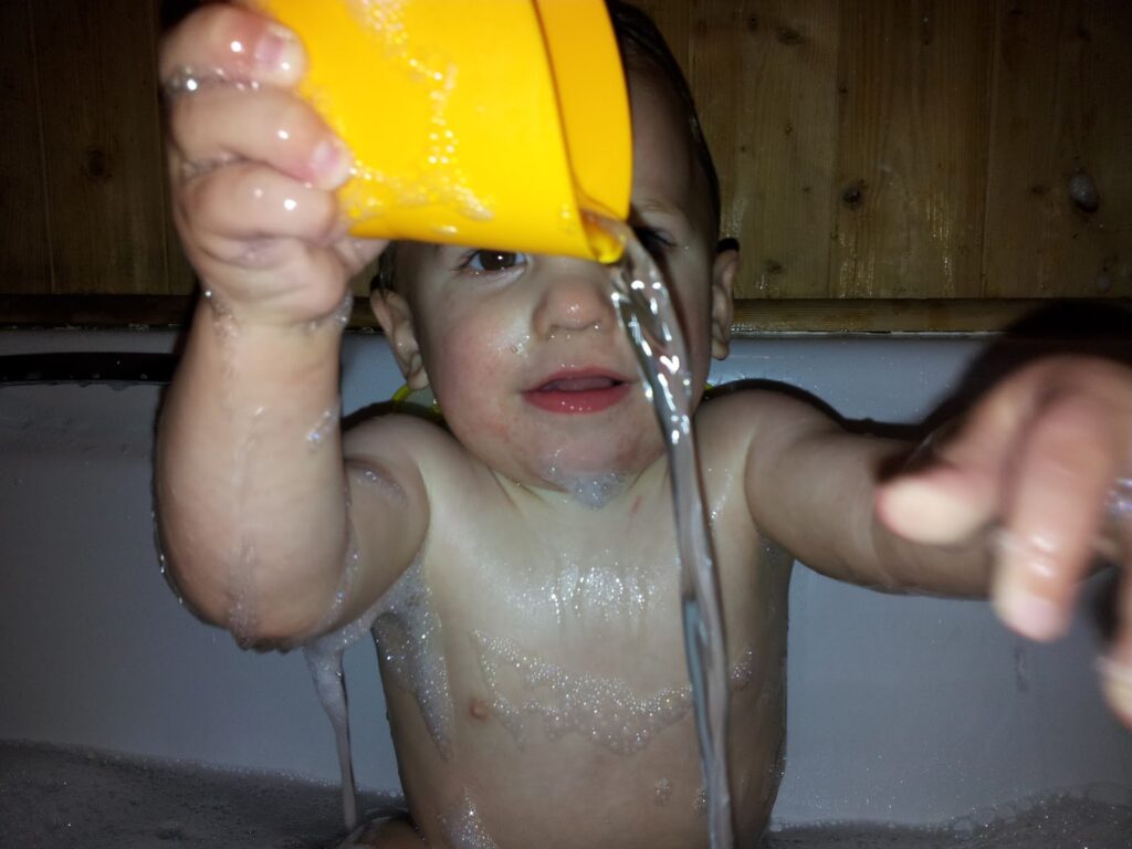 A young child sits in a bath, holding a yellow cup above their head and pouring water out. The child is wet, with some bubbles on their arms and chest, and reaches forward with their other hand. The background features wooden panelling.