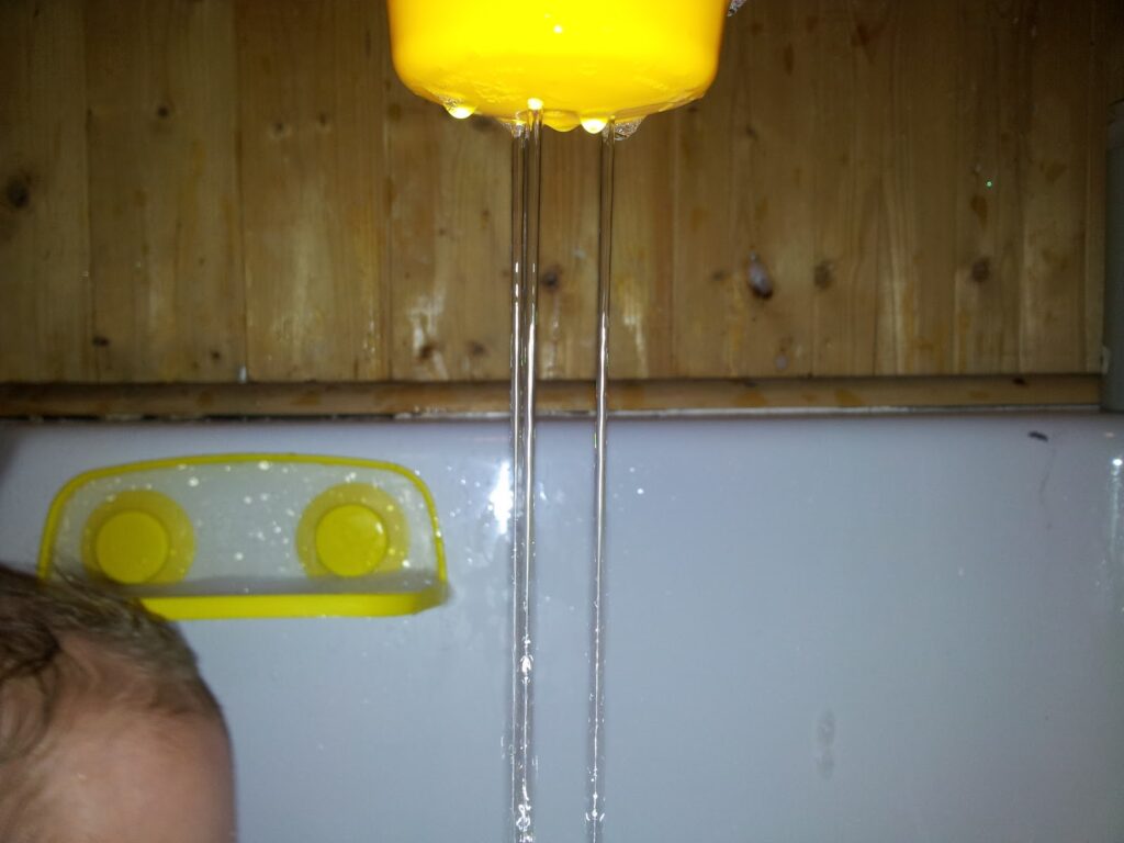 Close-up of water streaming from a yellow cup above a white bath. Part of a child's head is visible in the lower left corner. The bath has a yellow bath toy holder attached to the wall; the background is light wood panelling.