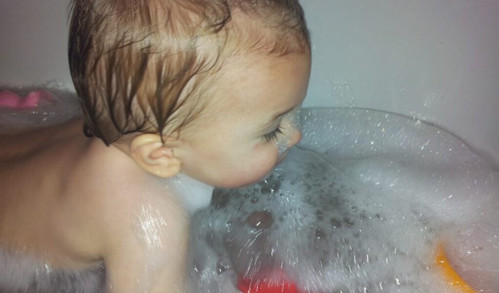 A baby with wet hair is lying in a bath filled with soapy bubbles, facing to the right. The child’s face and upper body are visible, and coloured bath toys float among the bubbles in the water. The setting appears playful and bright.