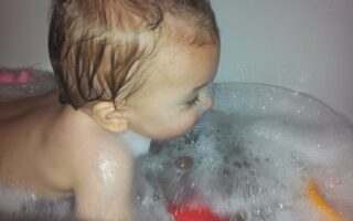 A baby with wet hair is lying in a bath filled with soapy bubbles, facing to the right. The child’s face and upper body are visible, and coloured bath toys float among the bubbles in the water. The setting appears playful and bright.