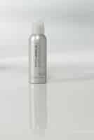 A silver spray bottle with a white cap stands upright on a glossy white surface. The label reads “paul mitchell” in lowercase letters. The background is plain and softly lit, creating a clean, minimalist look with a faint reflection below the bottle.