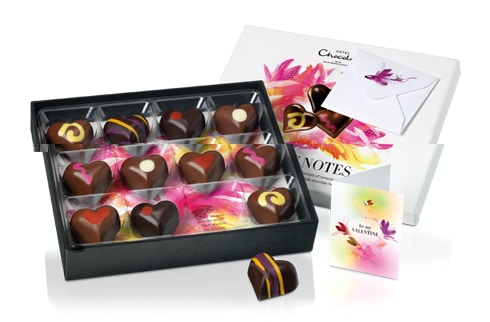 A box of assorted heart-shaped chocolates in various designs sits partly open. The packaging is white with pink and yellow floral artwork. Beside the box are a floral greetings card and an envelope, suggesting a gift for a special occasion.
