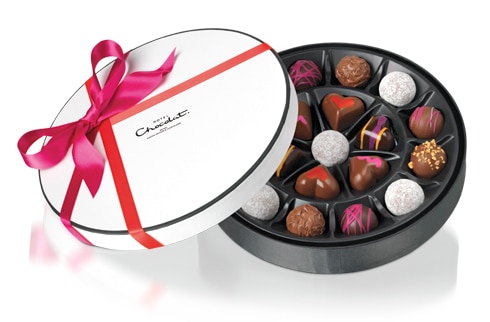 A round box of assorted chocolates with a white lid, red ribbon, and bow. The chocolates are various shapes and decorated with colourful patterns, including hearts, stripes, and sprinkles, arranged neatly in individual compartments inside the box.