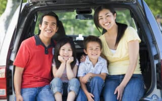A smiling family of four, with two adults and two children, sits close together in the open boot of a car. They are outdoors on a sunny day, casually dressed in jeans and shirts, appearing happy and ready for a day out or a trip.