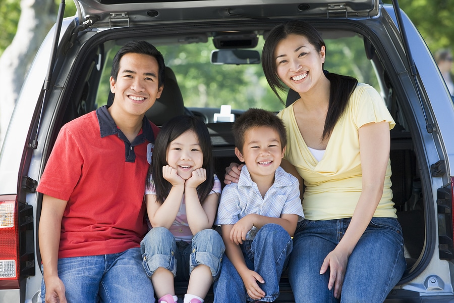 A smiling family of four, with two adults and two children, sits close together in the open boot of a car. They are outdoors on a sunny day, casually dressed in jeans and shirts, appearing happy and ready for a day out or a trip.
