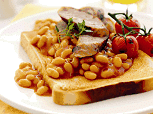 A plate with baked beans in tomato sauce on toasted bread, topped with slices of sausage and a sprig of herbs. Roasted cherry tomatoes are on the side, and the background shows a glass of orange juice.