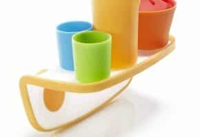 A set of four colourful plastic cups—yellow, orange, blue, and green—placed on a yellow and white tray shaped like a boat. The cups are of different sizes and arranged upright, with the tray balancing on a curved bottom.