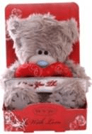 A fluffy grey teddy bear with a blue nose sits in a red box decorated with roses and cursive writing. The bear holds a red heart-shaped cushion, and the box reads “With Love” at the front. The bear appears soft and cuddly.