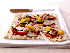 A rectangular flatbread pizza topped with tomato sauce, yellow peppers, mushrooms, and aubergine sits on a white textured cloth. A knife with a wooden handle rests behind the pizza, and one corner slice is slightly separated from the rest.