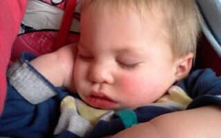 A young child with blond hair is sleeping with one hand near their face, resting on an adult’s lap. The child wears a striped shirt and appears peaceful. The adult, dressed in a red hoodie with white lettering, is partly visible, providing a comforting embrace.