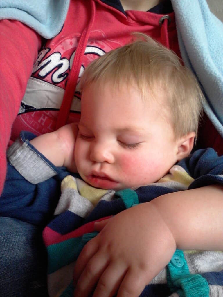 A young child with short blond hair sleeps peacefully, resting their head on one hand. The child wears a striped outfit with blue, green, and yellow colours. They are nestled against an adult in a red and blue hoodie, partially covered by a light blue blanket.