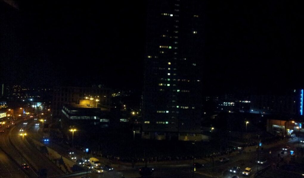 A cityscape at night shows a tall, dark building with illuminated windows. Streetlights and car headlights light up curving roads below, with several vehicles and some lit buildings visible in the background against the dark sky.