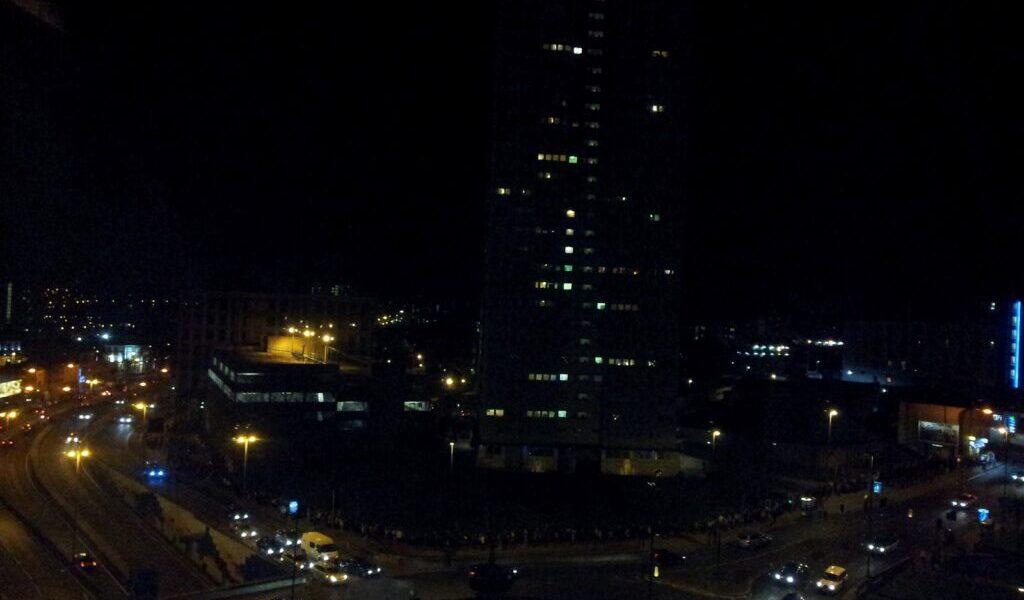A cityscape at night shows a tall, dark building with illuminated windows. Streetlights and car headlights light up curving roads below, with several vehicles and some lit buildings visible in the background against the dark sky.