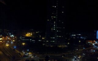 A cityscape at night shows a tall, dark building with illuminated windows. Streetlights and car headlights light up curving roads below, with several vehicles and some lit buildings visible in the background against the dark sky.