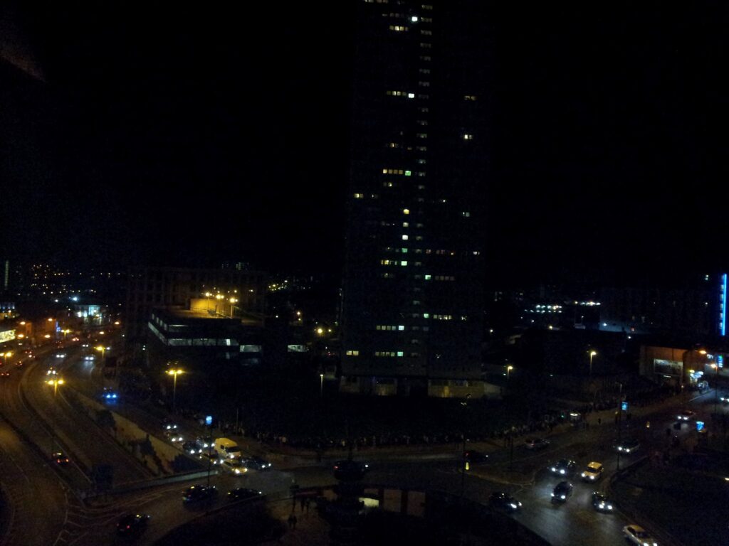 A tall, dark building stands in the centre of a city at night, faintly lit by scattered windows. Busy roads curve around the building, filled with moving cars and glowing streetlights. Surrounding buildings are dimly visible in the darkness.