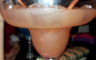 A large margarita glass filled with a pink frozen drink and ice, garnished with a white and an orange straw. Brightly coloured paper cocktail umbrellas and a sushi roll are visible in the background on a dark table. The image is slightly blurry.