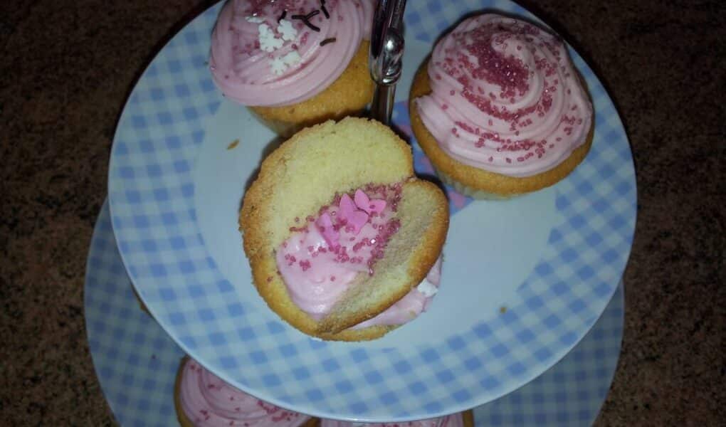 A two-tier, blue-and-white chequered cake stand holds cupcakes with pink icing and pink hundreds and thousands. One cupcake on the top tier is split in half, revealing more pink icing and hundreds and thousands inside. The stand is on a brown worktop.