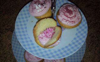 A two-tier, blue-and-white chequered cake stand holds cupcakes with pink icing and pink hundreds and thousands. One cupcake on the top tier is split in half, revealing more pink icing and hundreds and thousands inside. The stand is on a brown worktop.