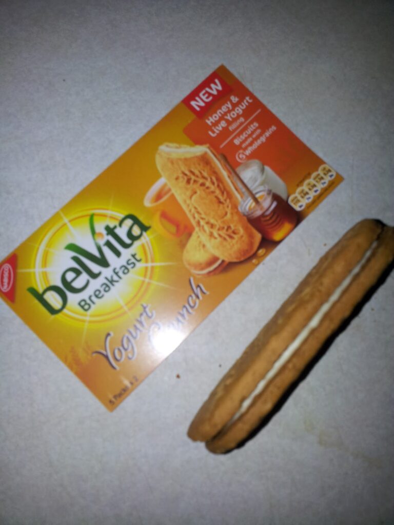 A box of belVita Breakfast Yoghurt Crunch biscuits, Honey & Live Yoghurt flavour, sits on a light surface. Next to the box, a single sandwich biscuit filled with white yoghurt cream is displayed, matching the image on the packaging.