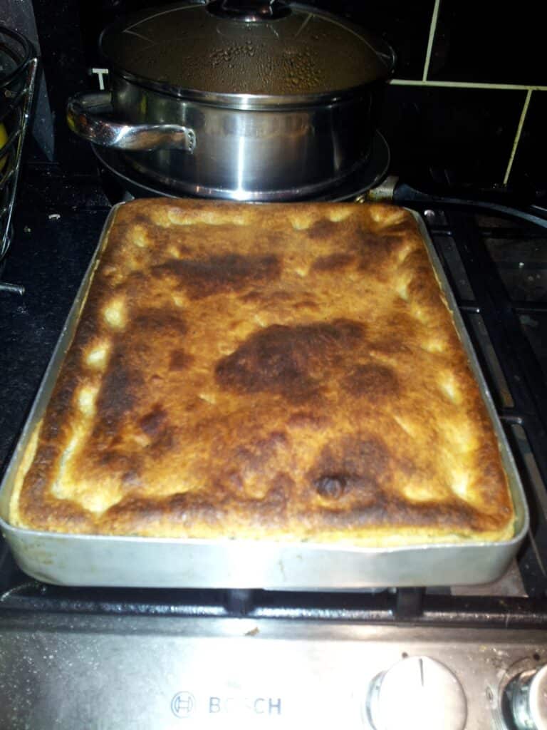 A large rectangular pie with a golden-brown, slightly overcooked pastry sits in a metal baking tray on a hob. In the background, there is a stainless steel saucepan with a lid and black kitchen tiles. The oven dial is visible in front.