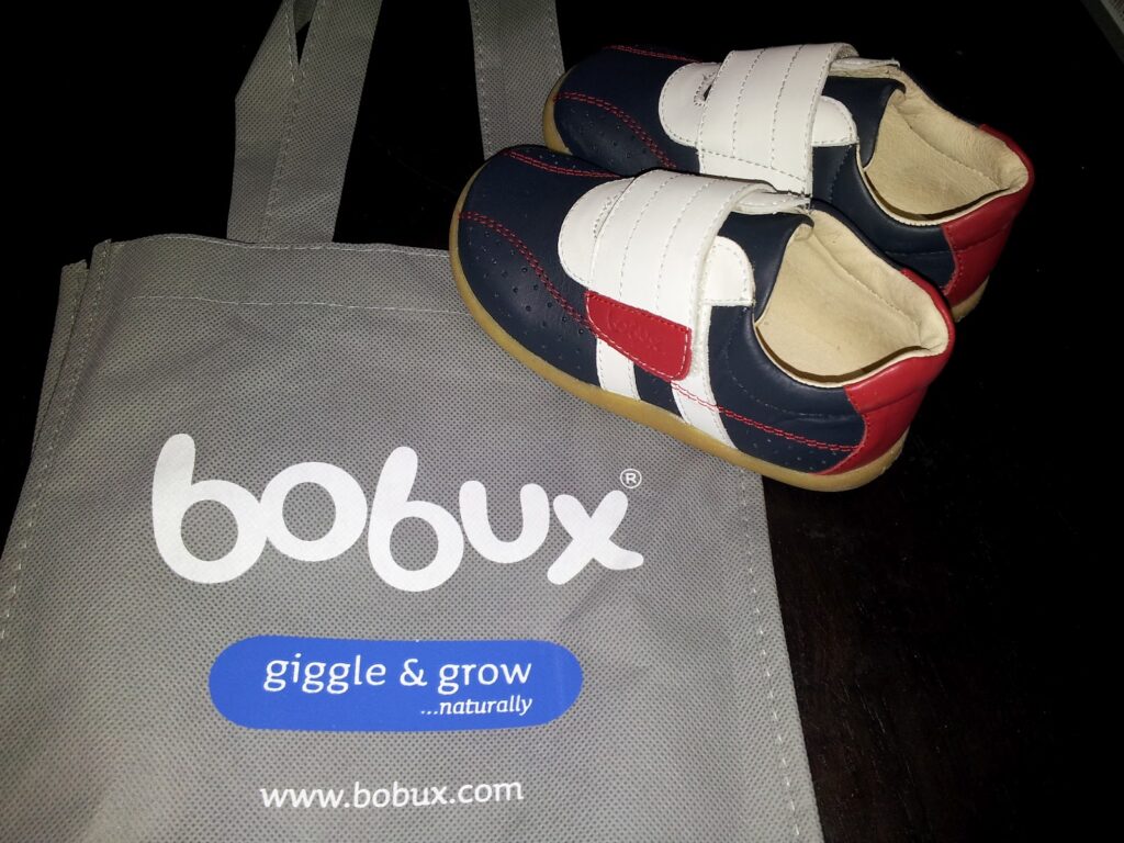 A pair of toddler shoes in navy blue, white, and red with Velcro straps sits on a grey Bobux tote bag that displays the white Bobux logo and the slogan “giggle & grow...naturally” above the website “www.bobux.com”.