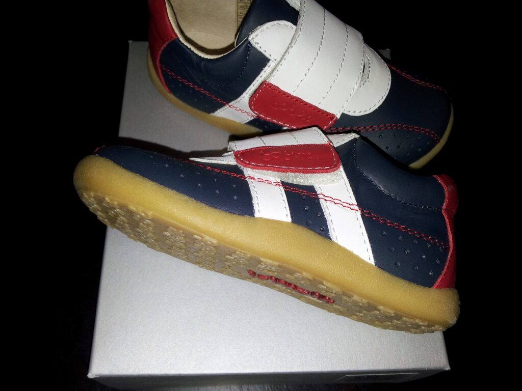 A pair of children’s trainers with navy blue, white, and red panels, Velcro straps, and tan rubber soles. The shoes are positioned on top of a white box, with one shoe resting on its side and the other standing upright. The background is black.