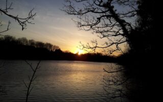 A serene lake at sunset with the sun low on the horizon, casting a golden glow over the water. Silhouetted tree branches frame the scene, and the sky transitions from blue to warm orange tones. The water reflects the soft light of dusk.