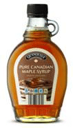 A clear glass bottle of pure Canadian maple syrup with a metal cap and a small handle, featuring a beige label with black and white text and an image of stacked pancakes topped with syrup. The syrup inside is amber coloured.